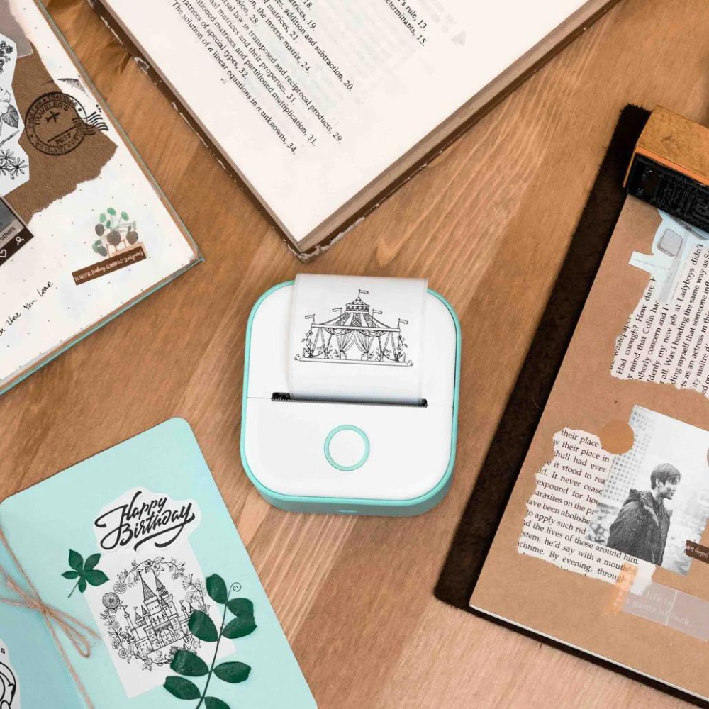 Stampante termica portatile bianca su tavolo di legno, circondata da quaderni e biglietti decorativi. Ideale per scrapbooking e stampa etichette.
