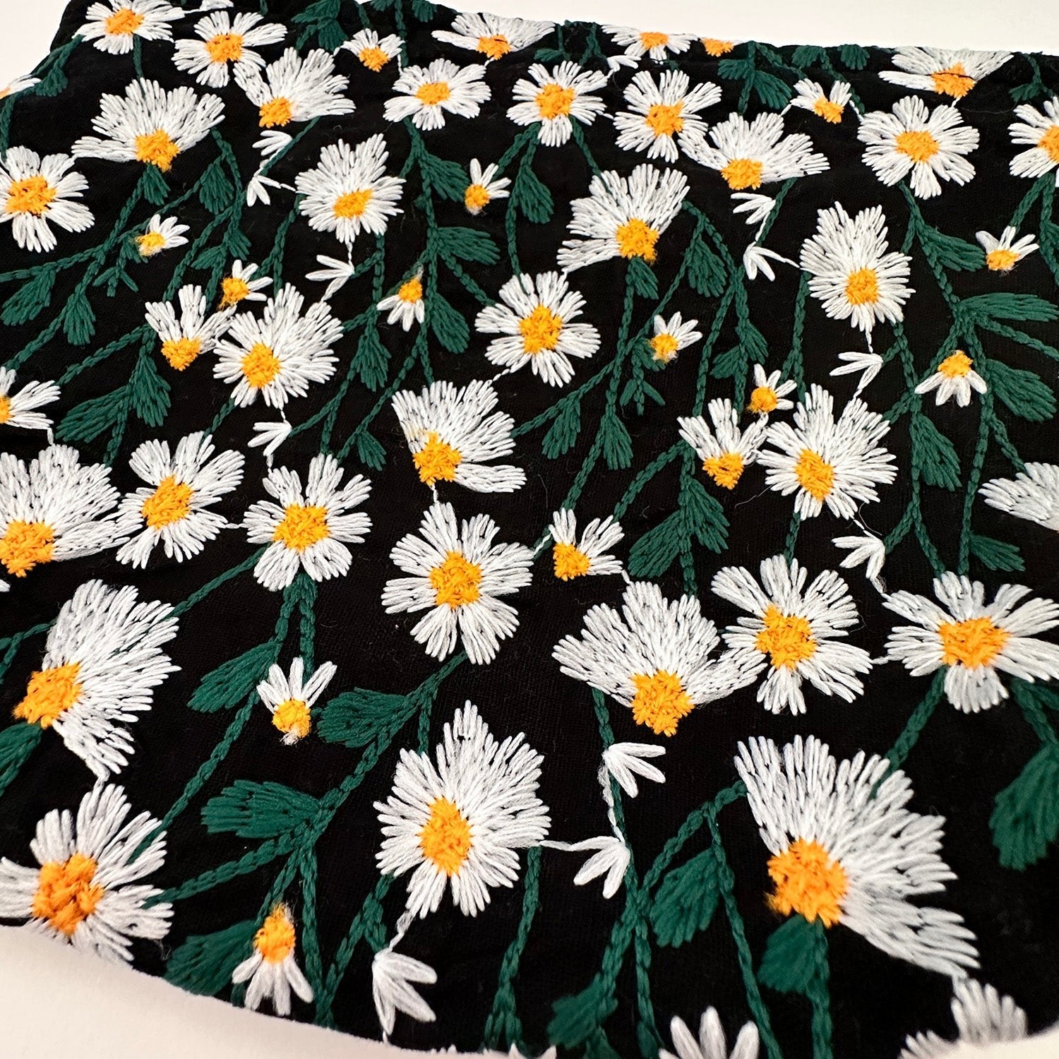 Tessuto nero ricamato con margherite bianche e gialle, motivo floreale, ideale per abbigliamento e decorazioni. Design elegante e vivace.