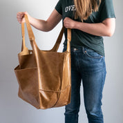 Donna con borsa shopper in pelle marrone, tasche laterali, indossa jeans e maglietta verde. Accessorio moda elegante e pratico per lo shopping.