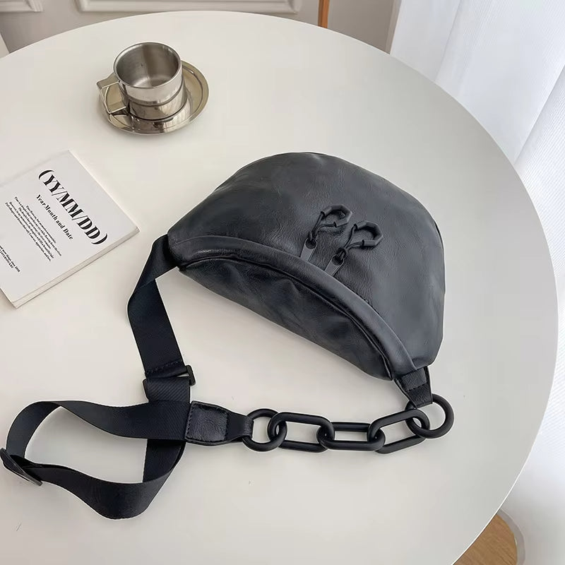 Marsupio nero in pelle su tavolo bianco, con catena decorativa e cinghia regolabile. Accessorio moda unisex, stile urbano e contemporaneo.