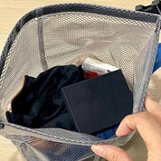 Borsa organizer in rete grigia con oggetti personali, tra cui un libro nero e vestiti. Ideale per viaggi, organizzazione bagagli, accessori viaggio.