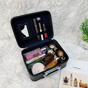 Valigetta cosmetica nera aperta su tappeto bianco, contenente profumi, rossetti, pennelli trucco e specchietto. Accanto, rivista di profumi e pianta decorativa.