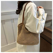 Donna con borsa a tracolla in paglia intrecciata, stile casual, manico in pelle marrone, indossa camicia bianca. Accessorio moda estivo elegante.