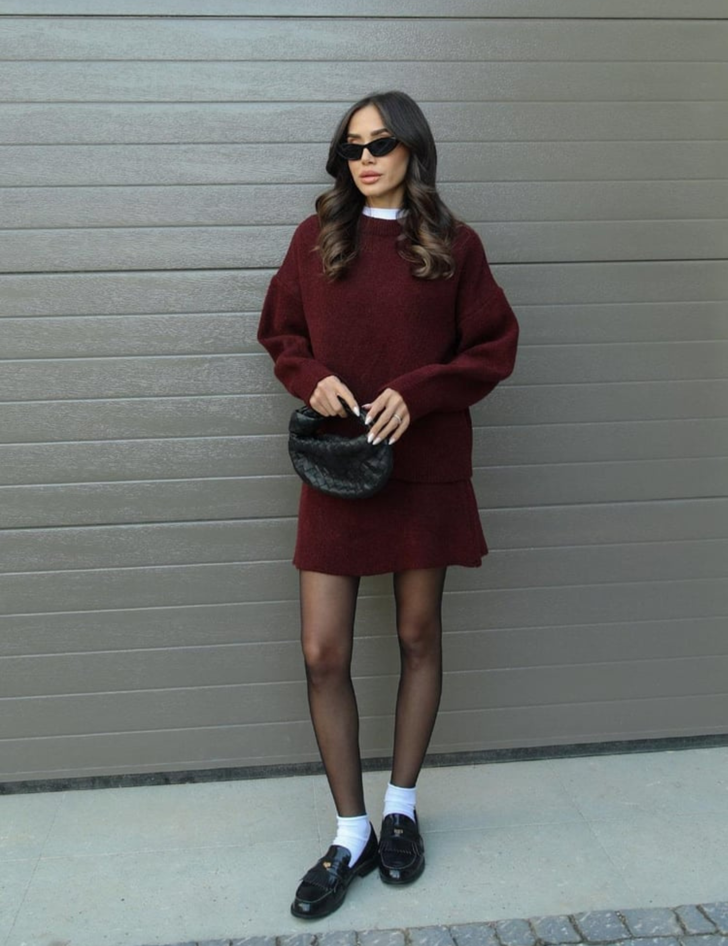 Donna con abito bordeaux, occhiali da sole neri, borsa a mano nera, calze nere e mocassini. Moda autunnale elegante e moderna.