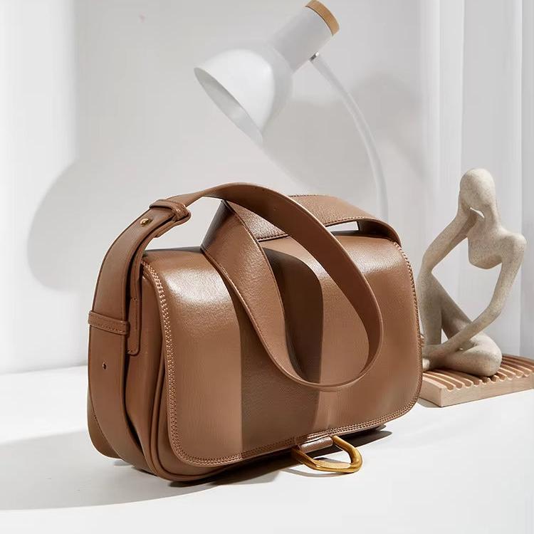Borsa a tracolla in pelle marrone su tavolo bianco, con lampada moderna e scultura astratta. Accessorio moda elegante e versatile.