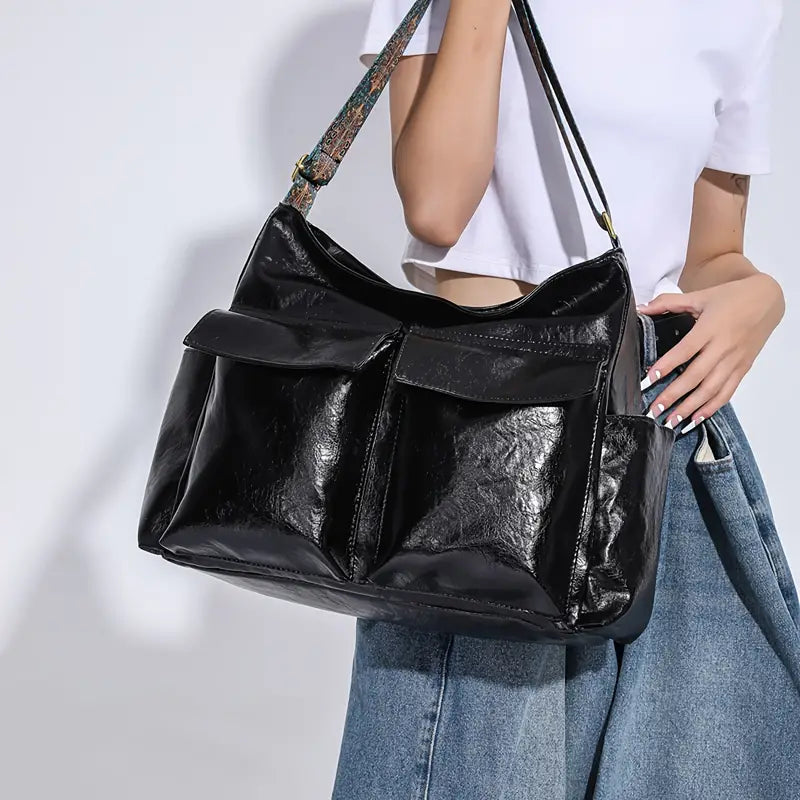Borsa a tracolla nera in pelle lucida, tasche frontali, indossata da donna con jeans e maglietta bianca. Accessorio moda elegante e spazioso.