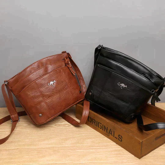 Due borse a tracolla in pelle, una marrone e una nera, su sfondo grigio. Design elegante con logo argentato, ideali per moda uomo e donna.