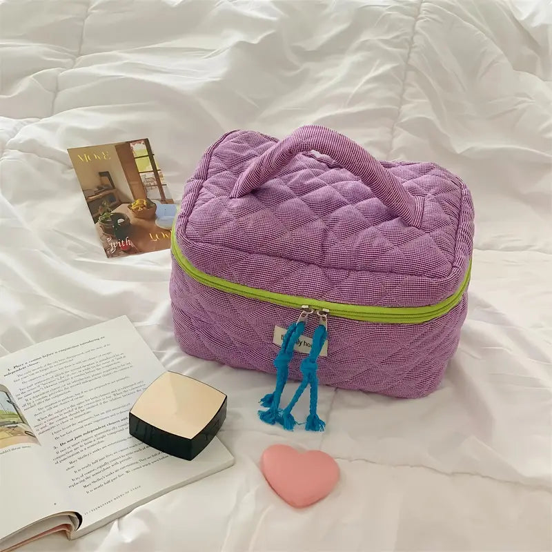 Borsa cosmetica viola trapuntata su letto bianco, con cerniera verde e nappine blu, accanto a libro aperto e cuore rosa. Accessori da viaggio eleganti.