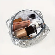 Borsa cosmetica argento aperta con trucchi: fondotinta, palette ombretti, flaconi skincare. Accessori bellezza, organizzazione trucco viaggio.