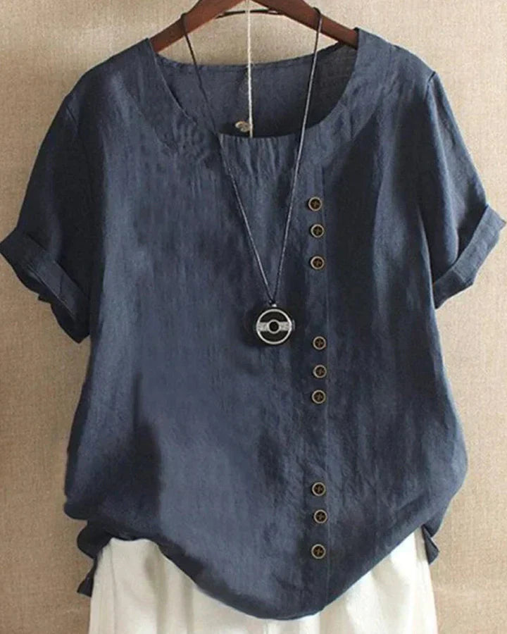 Maglietta donna in lino blu scuro con bottoni decorativi asimmetrici, maniche corte e collana pendente, appesa su gruccia.