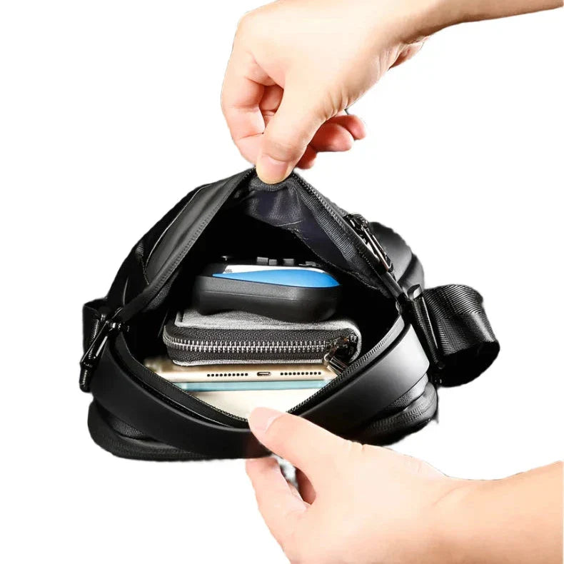 Mani che aprono una borsa a tracolla nera, mostrando smartphone, portafoglio e chiavi all'interno. Accessorio moda uomo, pratico e spazioso.