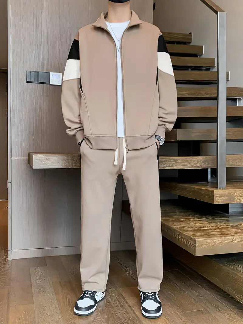 Tuta sportiva beige da uomo con giacca a zip e pantaloni abbinati, dettagli bianchi e neri, indossata con sneakers bianche e nere su pavimento in legno.