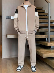 Tuta sportiva beige da uomo con giacca a zip e pantaloni abbinati, dettagli bianchi e neri, indossata con sneakers bianche e nere su pavimento in legno.