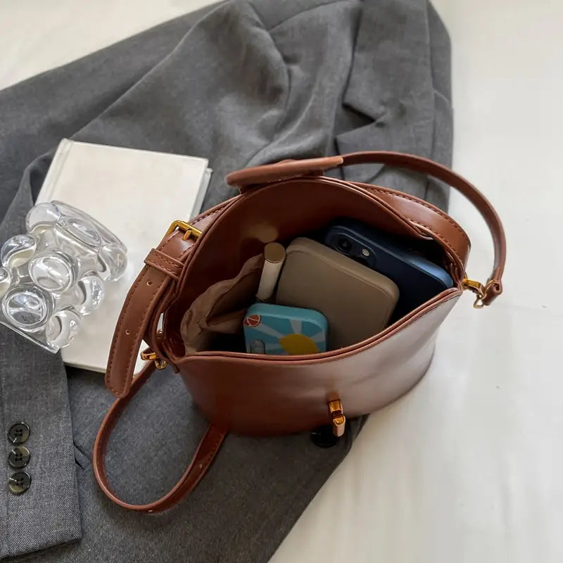 Borsa a secchiello in pelle marrone su giacca grigia, contenente smartphone, portafoglio, rossetto e specchio. Accessori moda eleganti e pratici.