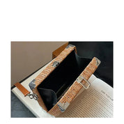 Borsa a tracolla in paglia intrecciata con dettagli in metallo, design rettangolare, aperta su libro, stile estivo elegante e moderno.