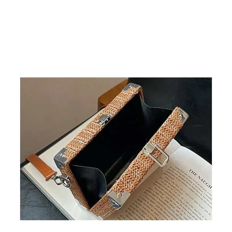 Borsa a tracolla in paglia intrecciata con dettagli in metallo, design rettangolare, aperta su libro, stile estivo elegante e moderno.