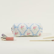 Astuccio trapuntato bianco con fiocchi blu e rose rosa, circondato da cosmetici e pennelli. Accessorio elegante per trucco e bellezza.