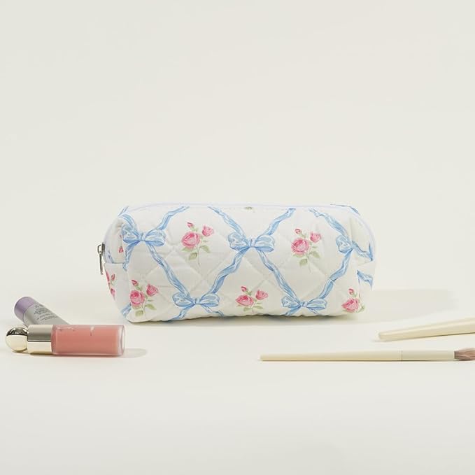 Astuccio trapuntato bianco con fiocchi blu e rose rosa, circondato da cosmetici e pennelli. Accessorio elegante per trucco e bellezza.
