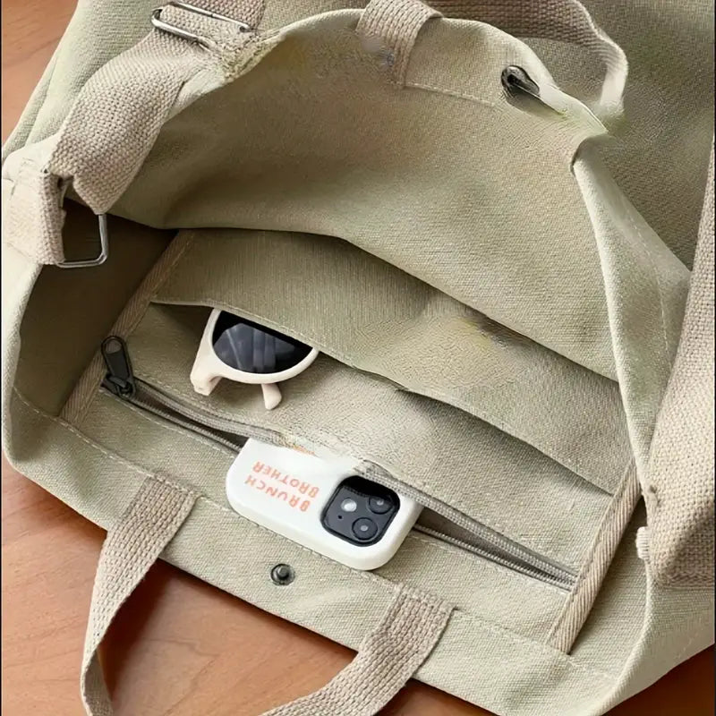 Borsa in tela beige aperta su tavolo di legno, contenente occhiali da sole bianchi e smartphone con custodia bianca. Accessori moda casual.