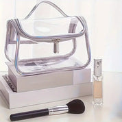 Borsa trasparente in PVC con cerniera, posizionata su libri, accanto a pennello da trucco e flacone di profumo. Accessori moda e bellezza.