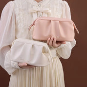 Donna in abito bianco tiene due pochette in pelle, una rosa e una bianca, su sfondo marrone. Elegante accessorio moda femminile.