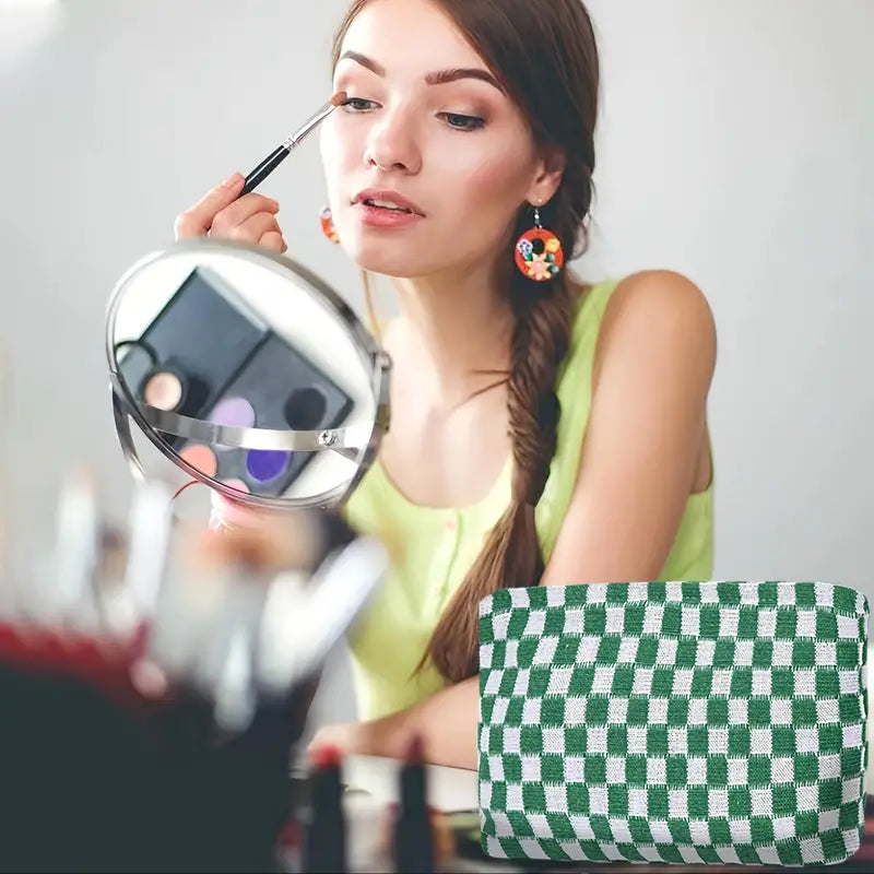 Giovane donna si trucca davanti a uno specchio, usando un pennello per ombretto. Borsa a scacchi verde e bianca in primo piano.