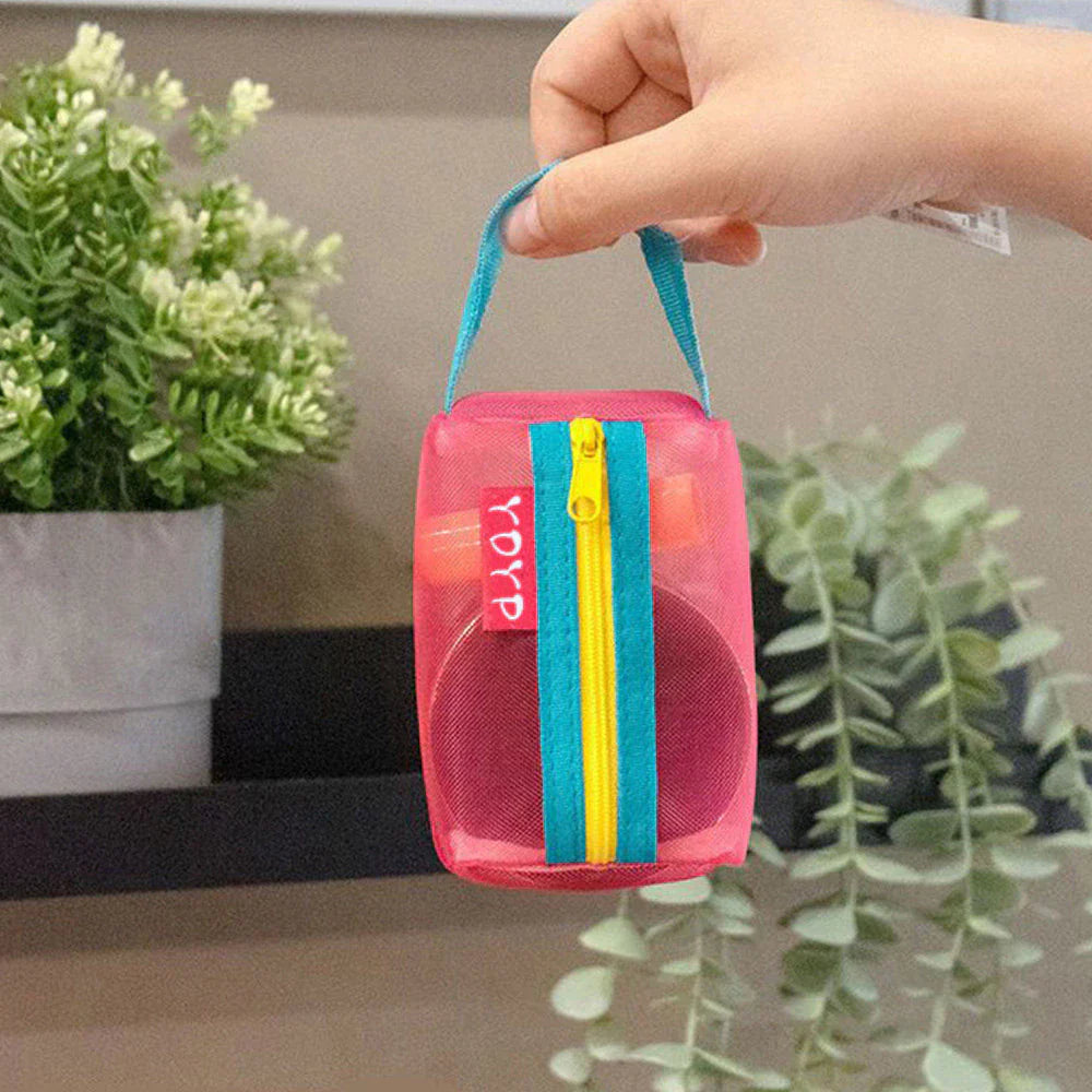 Mano che tiene una borsa trasparente rosa con cerniera gialla e blu, contenente un oggetto rotondo. Sfondo con piante decorative.