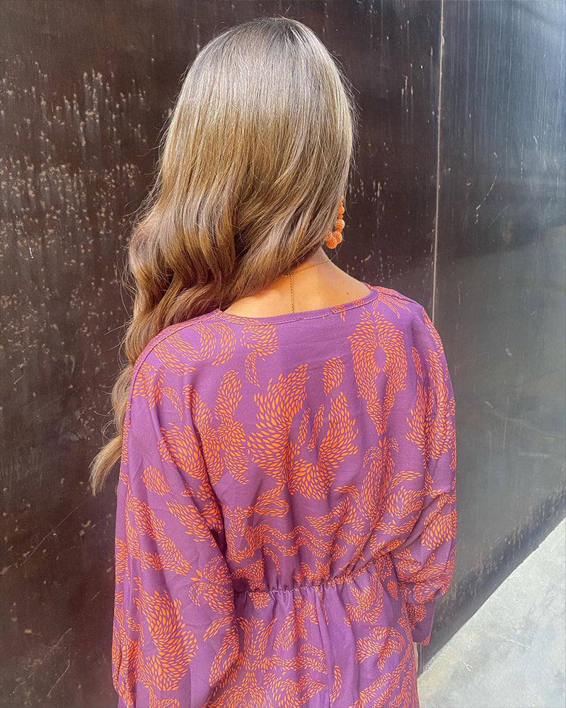 Donna di spalle con capelli ondulati, indossa abito viola con motivi arancioni. Moda autunnale, abbigliamento femminile elegante, tendenze 2023.