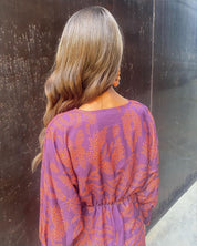 Donna di spalle con capelli ondulati, indossa abito viola con motivi arancioni. Moda autunnale, abbigliamento femminile elegante, tendenze 2023.
