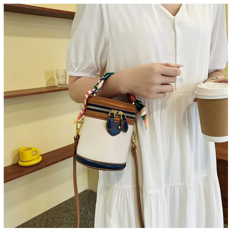 Donna con abito bianco tiene borsa a tracolla cilindrica beige e blu, decorata con foulard colorato, e bicchiere di caffè. Accessori moda eleganti.
