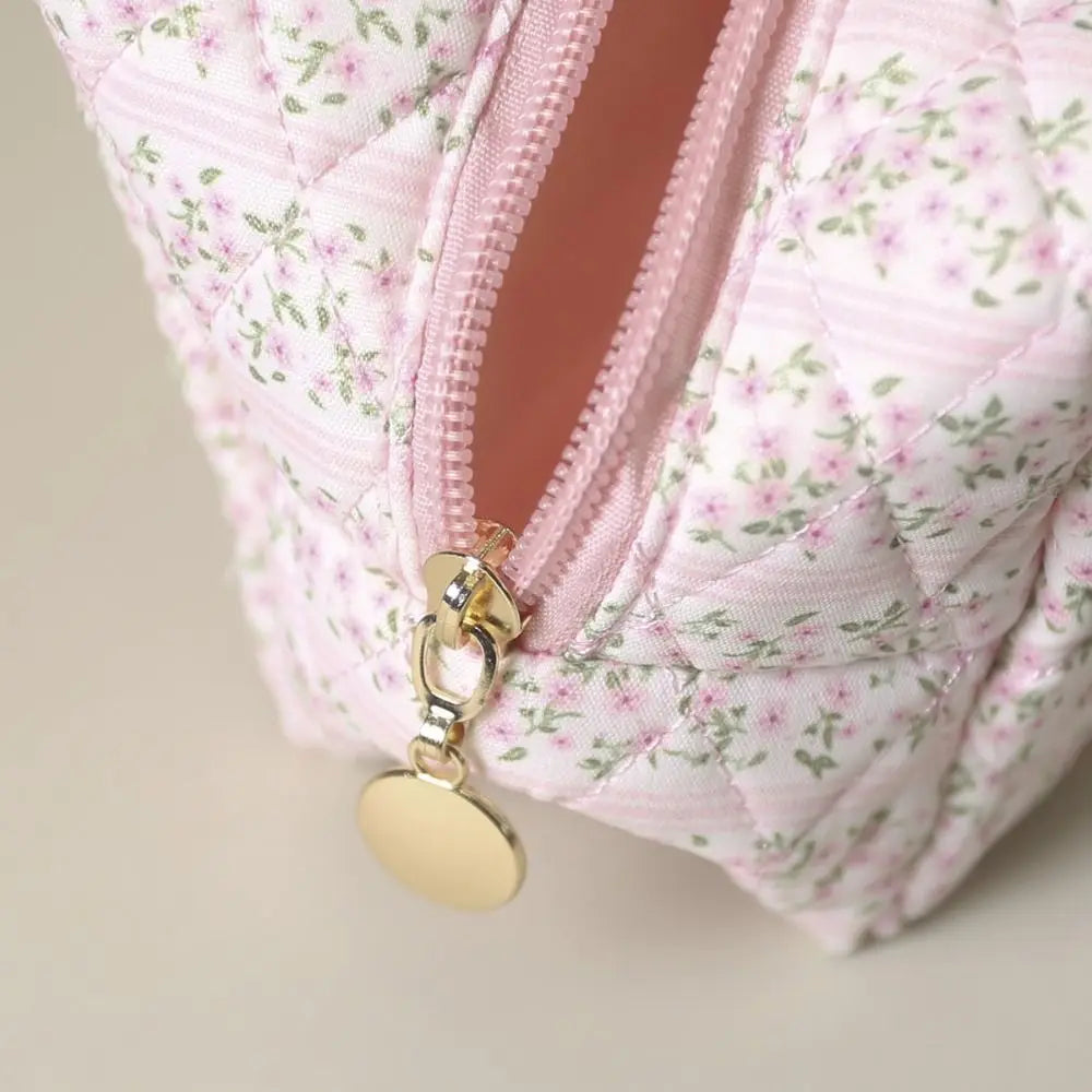 Astuccio cosmetico trapuntato rosa con motivo floreale, cerniera dorata e pendente rotondo. Accessorio elegante per trucco e viaggio.