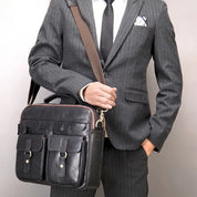 Uomo in abito grigio gessato con cravatta nera, tiene una borsa a tracolla in pelle nera con tasche frontali. Elegante accessorio moda business.