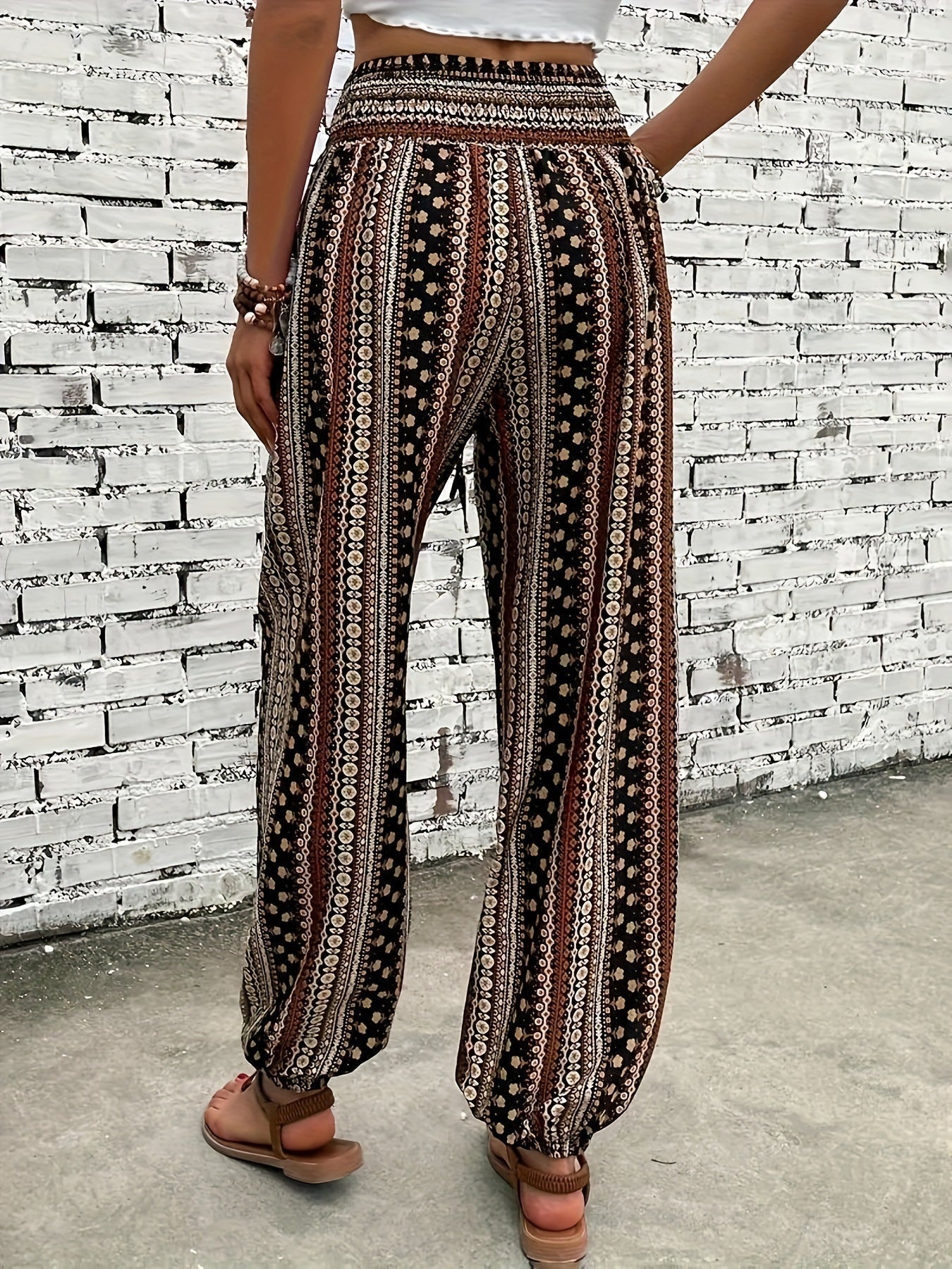 Pantaloni palazzo donna, motivo etnico, colori marrone e bianco, tessuto leggero, indossati con sandali, sfondo muro bianco.