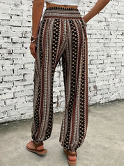 Pantaloni palazzo donna, motivo etnico, colori marrone e bianco, tessuto leggero, indossati con sandali, sfondo muro bianco.