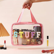 Borsa trasparente con manici rosa, contenente cosmetici e accessori, decorata con lettere colorate "STUFF". Ideale per organizzare trucchi e prodotti di bellezza.