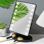 Specchio cosmetico LED rettangolare con foglia verde riflessa, base nera, circondato da pennelli trucco e pianta decorativa. Perfetto per make-up.