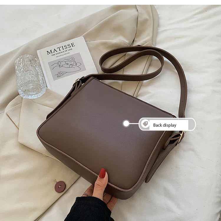 Borsa a tracolla marrone su tessuto beige, con libro d'arte e bicchiere. Design elegante, ideale per moda femminile e accessori di tendenza.