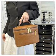 Donna con borsa a mano in pelle marrone, chiusura a zip dorata, indossa giacca nera e gonna bianca. Design elegante, accessorio moda femminile.