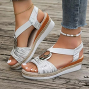 Sandali estivi bianchi con zeppa, cinturino alla caviglia, dettagli metallici, indossati su pavimento in legno. Scarpe moda donna casual eleganti.