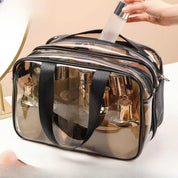 Borsa trasparente per cosmetici con manici neri, contenente flaconi di profumo e lozioni. Ideale per viaggi, organizzazione trucco, accessori beauty.