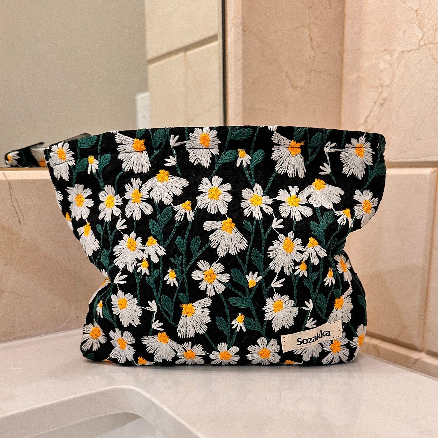 Borsa cosmetica nera con motivo floreale margherite bianche e gialle, marchio Sozakka, su sfondo bagno. Accessorio moda donna elegante e pratico.