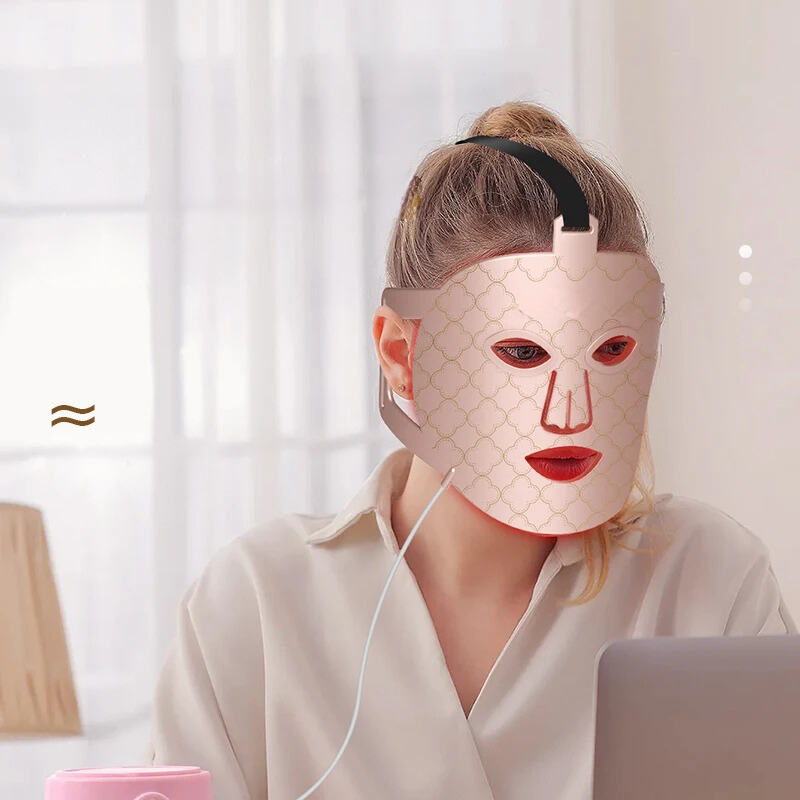 Donna con maschera LED per terapia della pelle, seduta al computer. Maschera viso bellezza, cura della pelle, trattamento anti-età, tecnologia LED.
