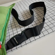 Cintura nera regolabile con fibbia in plastica su sfondo a griglia bianca e nera, accanto a tessuto verde con cerniera. Accessorio moda unisex.