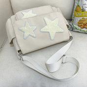 Borsa a tracolla beige con stelle bianche in rilievo, design moderno, su divano bianco. Accanto, confezione di snack al limone.