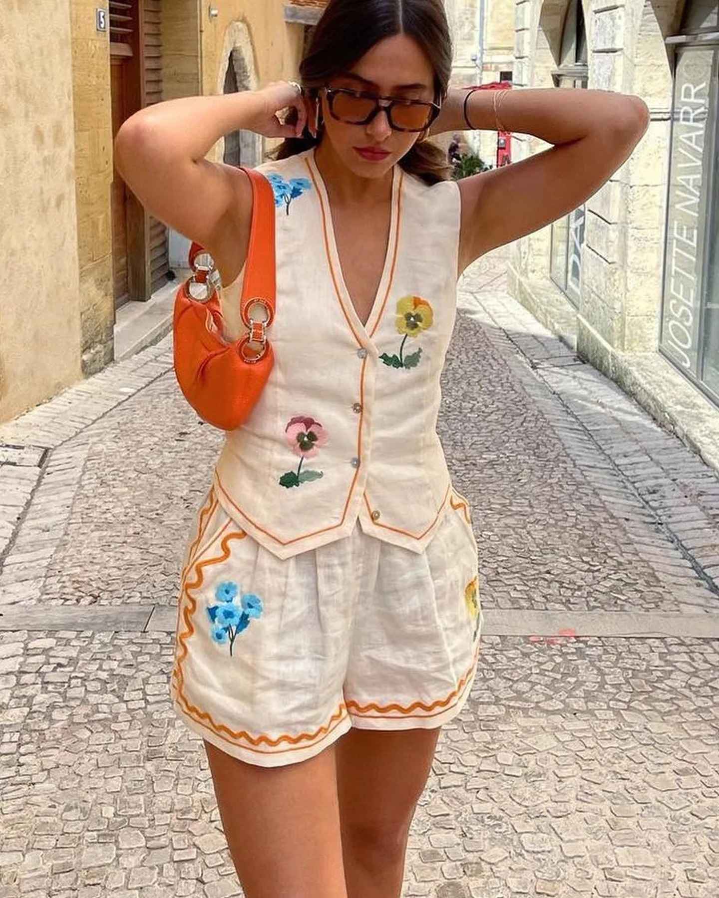 Donna con completo estivo bianco ricamato, borsa arancione, occhiali da sole, in strada acciottolata. Moda estiva, abbigliamento casual elegante.