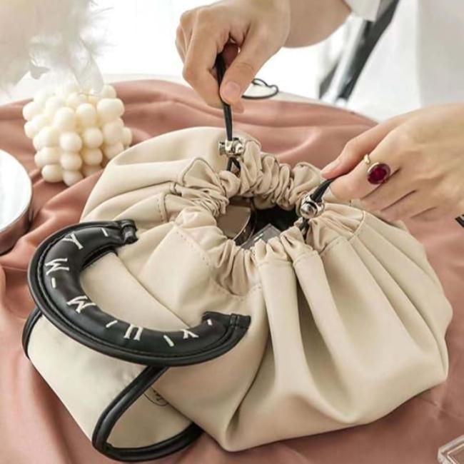 Borsa a secchiello beige con manico nero, chiusura a coulisse, su tavolo con tessuto rosa. Accessorio moda elegante, design moderno.