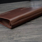 Custodia in pelle marrone per laptop su tavolo in legno, design elegante e sottile, ideale per protezione e trasporto sicuro di dispositivi elettronici.