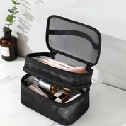 Borsa cosmetica nera a due scomparti aperti, contenente pennelli trucco e prodotti skincare, su sfondo bianco con pianta decorativa.