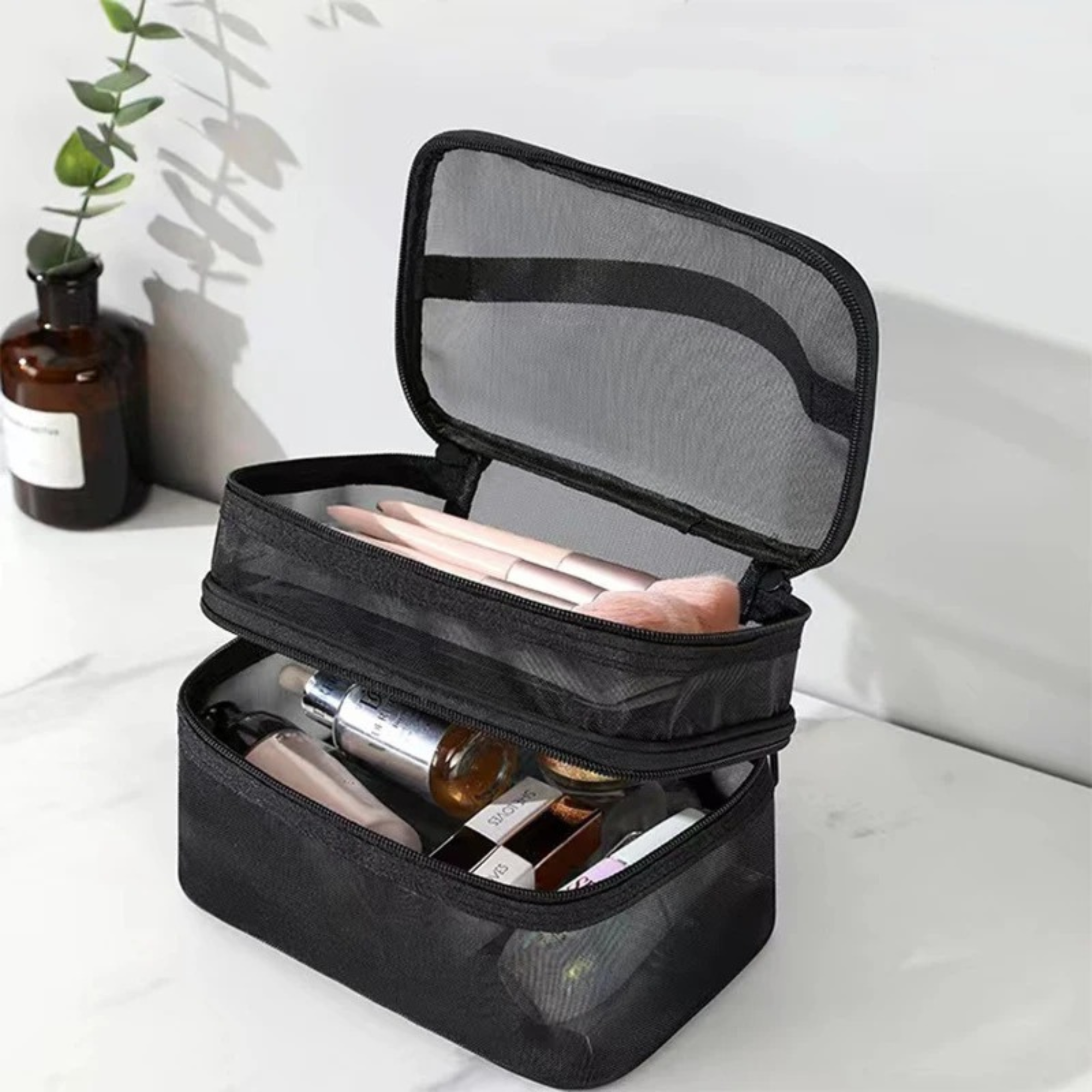 Borsa cosmetica nera a due scomparti aperti, contenente pennelli trucco e prodotti skincare, su sfondo bianco con pianta decorativa.