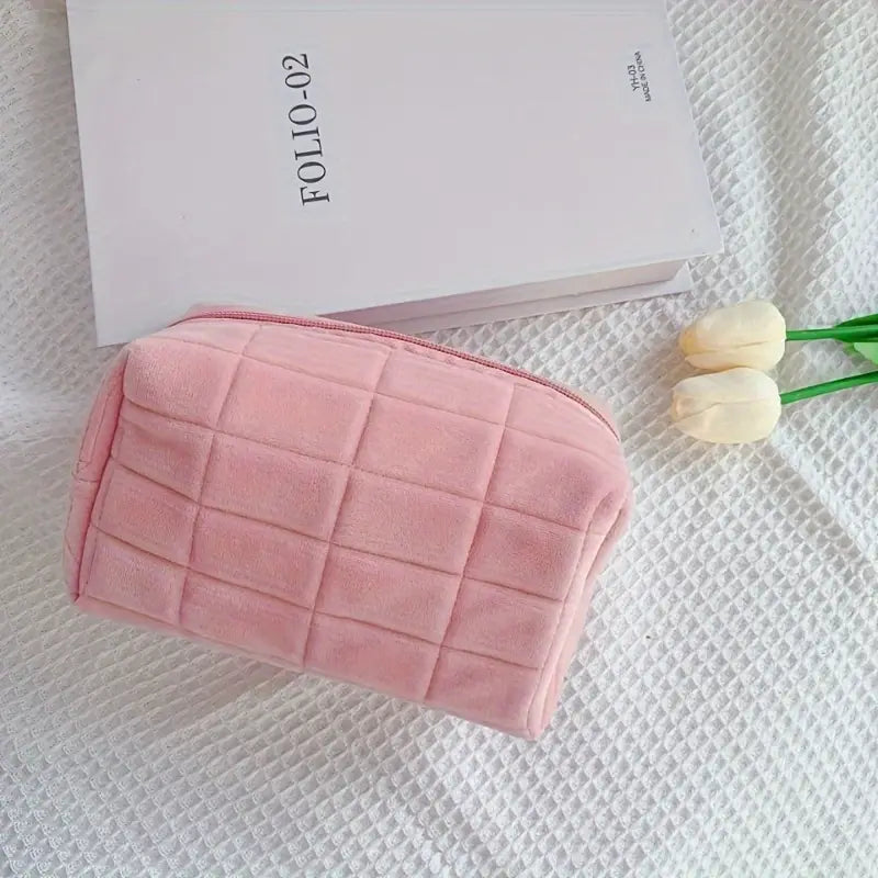 Astuccio cosmetico rosa in velluto su coperta bianca, accanto a libro e tulipani. Elegante e pratico, ideale per trucco e accessori da viaggio.